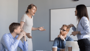 Foto de quatro pessoas, com roupas profissionais em volta de uma mesa. Duas sentadas e duas em pé. Representa conflitos no trabalho.