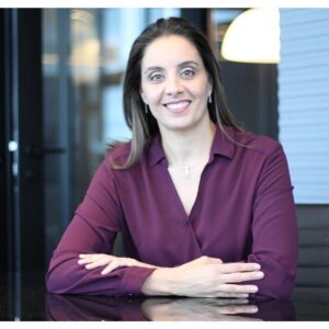 Foto de mulher branca com cabelos castanhos. Ela usa uma camisa violeta de manga longa e está sentada com os braços cruzados em cima de uma mesa. Apenas vemos do apoio da mesa para cima. Representa a entrevistada do artigo "O que faz um mediador extrajudicial?"
