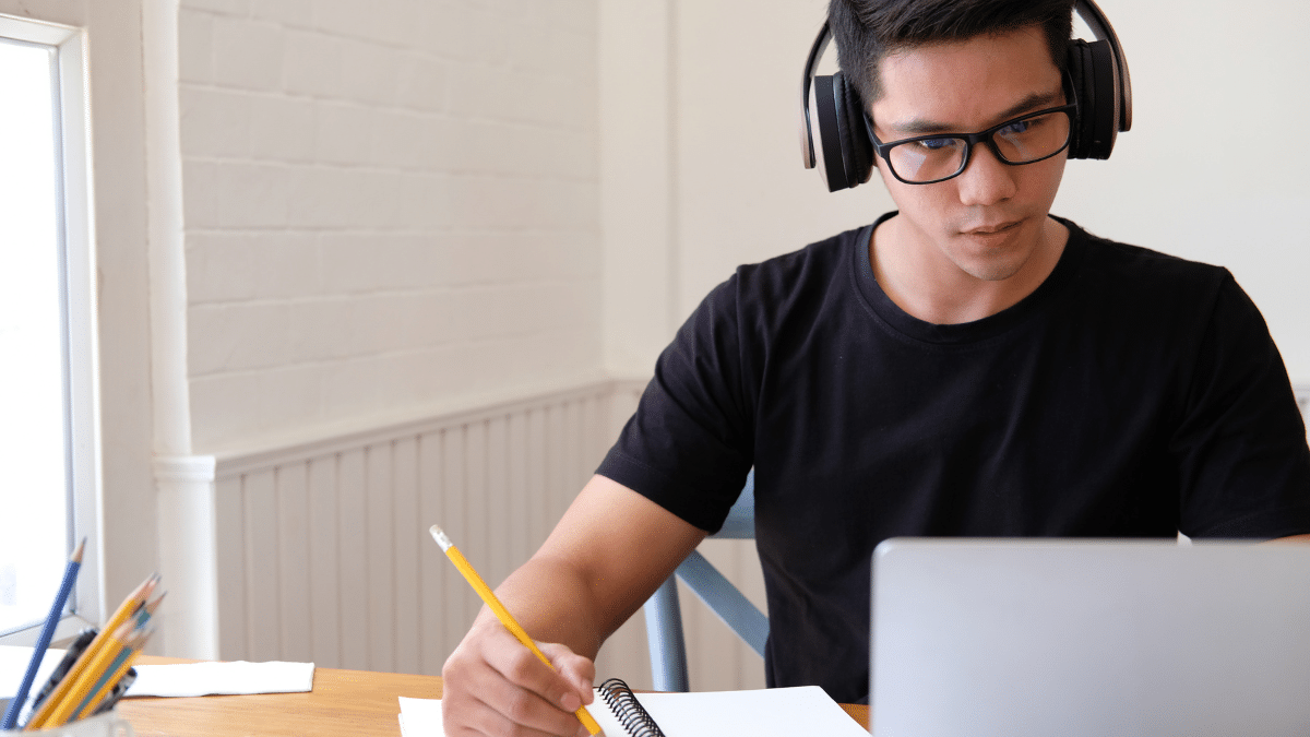 Homem estudando com fones de ouvido, lápis e caderno ao lado de um laptop, simbolizando aprendizado online e concentração.