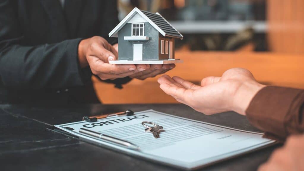 Foto de pessoas na mesa, foco nas suas mãos. Elas passam uma casa em miniatura. Representa justiça arbitral: posso perder meus bens?