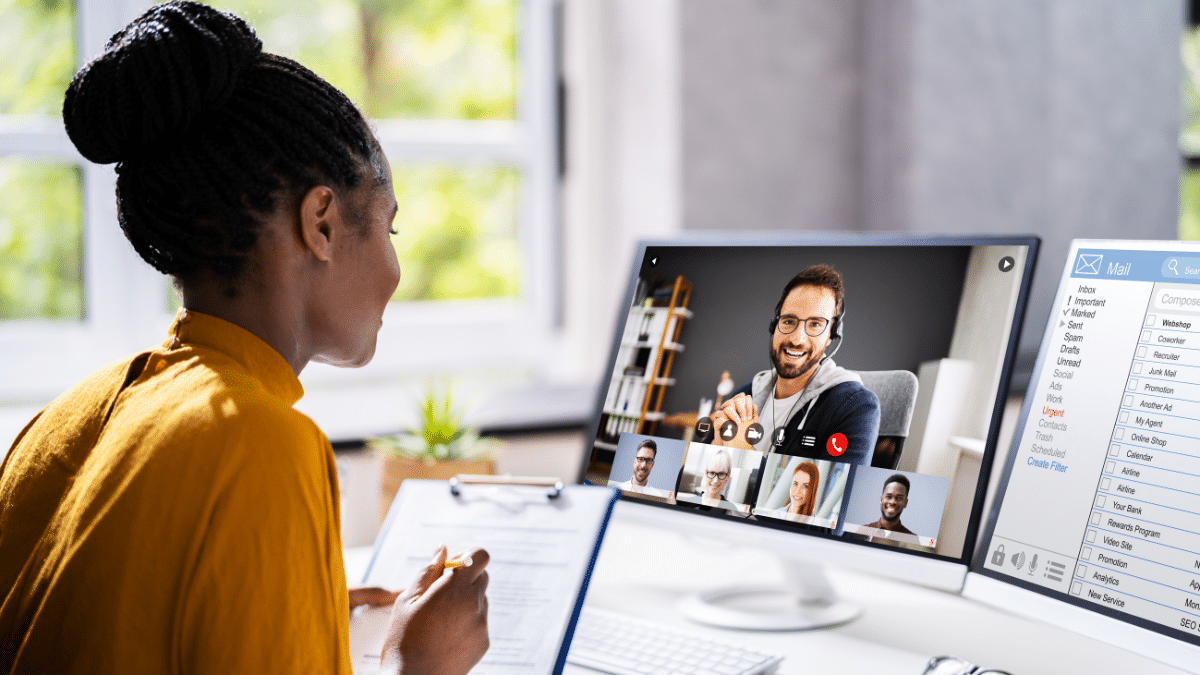 Mulher sentada à mesa, vestindo blusa mostarda, participando de uma videoconferência exibida em seu monitor. Na tela, um homem sorridente com fone de ouvido lidera a chamada, acompanhado por outros participantes em miniaturas. Ao lado, outro monitor exibe uma caixa de e-mail aberta. A imagem transmite profissionalismo, tecnologia e colaboração remota. Representa Audiência online.