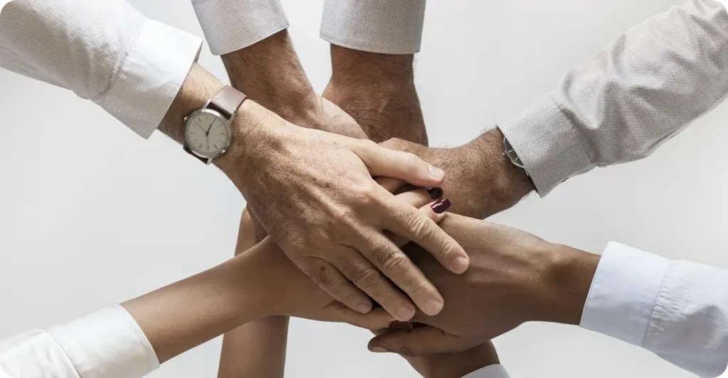 Imagem de várias mãos sobrepostas, simbolizando união, colaboração e trabalho em equipe. As mãos pertencem a diferentes pessoas, todas usando roupas formais, sugerindo um ambiente profissional e um compromisso coletivo.