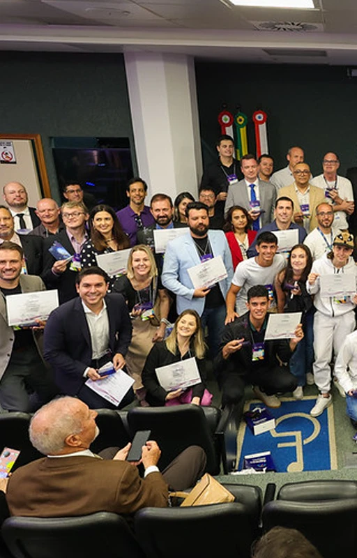 Um grande grupo de pessoas posando para uma foto coletiva em um auditório, com certificados e prêmios nas mãos. A imagem está relacionada ao "Prêmio de Pesquisa e Inovação Catarinense", com participantes sorridentes celebrando a ocasião.