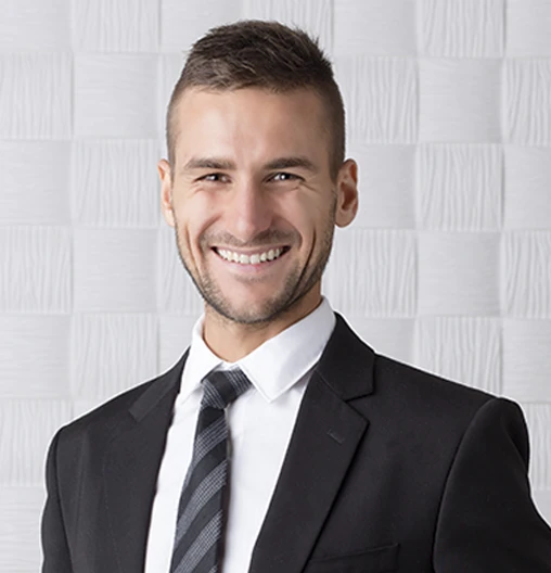 Homem jovem de terno sorrindo. Ele tem cabelo castanho e o fundo é branco.