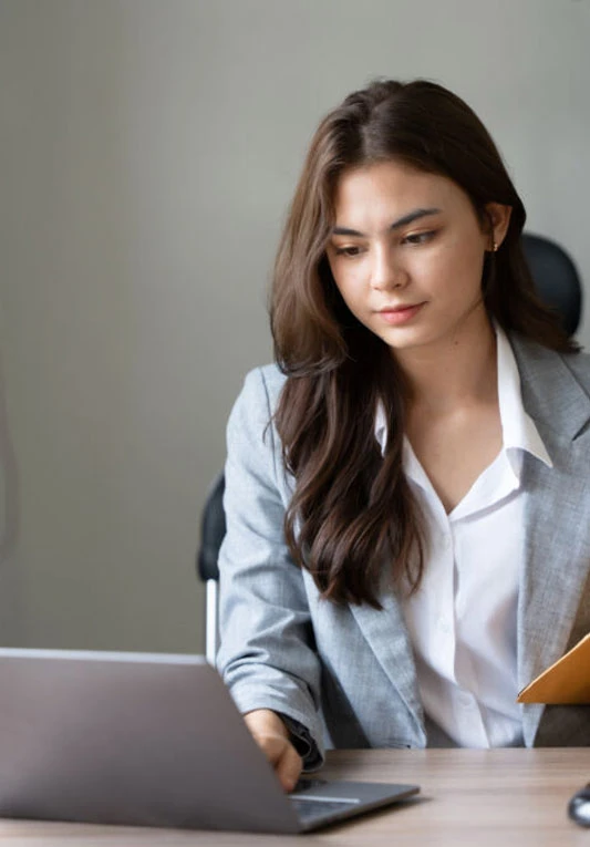 Imagem de mulher mexendo no computador. Ela tem pele clara e cabelo castanho escuro. Ela está sentada em uma mesa e parece focada.